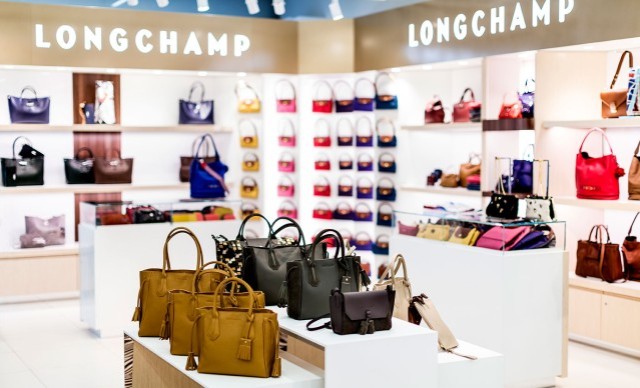 Longchamp-Geschäft mit verschiedenen Handtaschen auf weißen Tischen und Regalen, beleuchtet durch helle Lichter