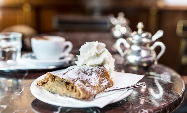 Apfelstrudel mit Puderzucker und Sahne auf einem Teller, im Hintergrund eine Tasse Kaffee und eine Teekanne.