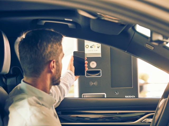 Ein Mann sitzt in einem Auto und hält sein Smartphone an eine Schrankenanlage, um kontaktlos zu bezahlen oder ein Parkticket zu scannen. Die Szene zeigt ein System von SKIDATA.