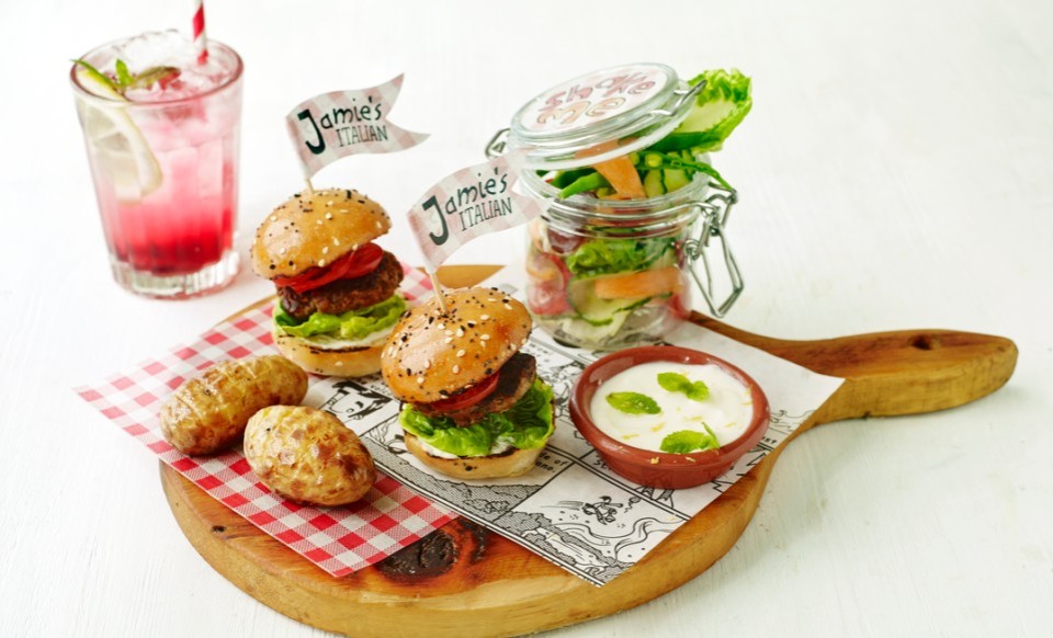 Zwei Mini-Burger mit "Jamie's Italian"-Fähnchen, Salat im Glas, zwei Würstchen und ein Getränk auf einem Holzbrett .