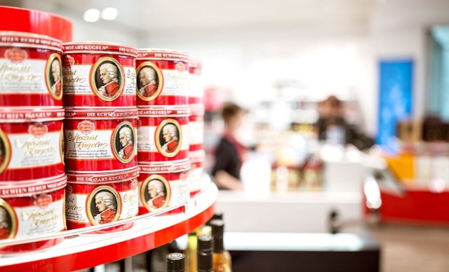 Rote Dosen mit Mozartkugeln in einem Geschäft, im Hintergrund unscharfe Personen und Regale.