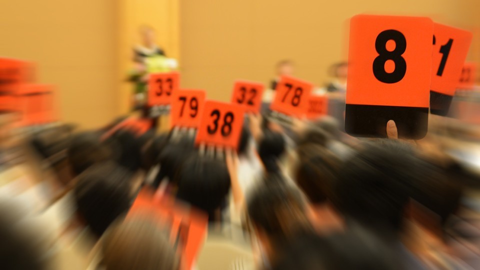 Menschen in einem Auktionssaal heben orangefarbene Bietertafeln mit schwarzen Zahlen, um Gebote abzugeben. Im Vordergrund ist die Tafel mit der Nummer 8 scharf zu sehen, während der Hintergrund verschwommen ist.