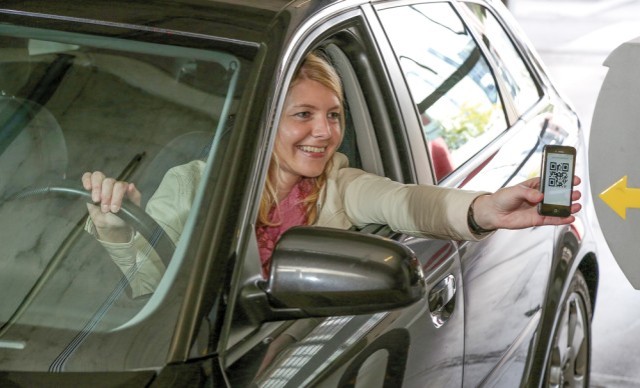 Frau im Auto hält ihr Smartphone mit QR-Code an einen Scanner am Parkhaus-Eingang.