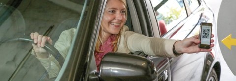 Frau im Auto hält ihr Smartphone mit QR-Code an einen Scanner am Parkhaus-Eingang.
