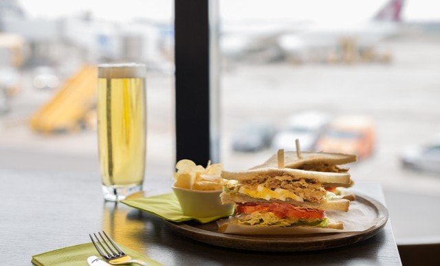 Sandwich mit Pommes und einem Glas Bier auf einem Tisch in einem Flughafenrestaurant, im Hintergrund unscharfe Flugzeuge