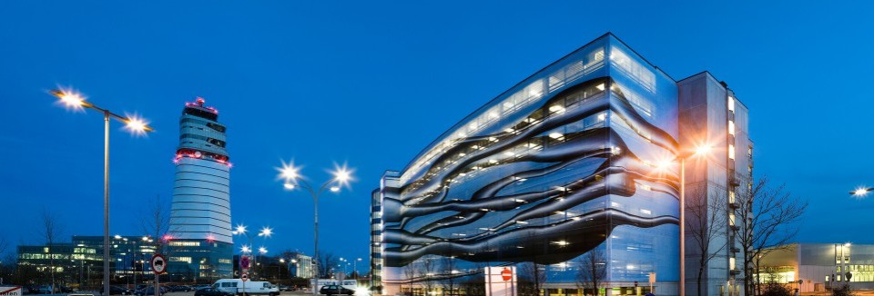 Beleuchtetes Parkhaus 3 mit moderner Fassade am Flughafen Wien bei Nacht, links der Tower mit roter Beleuchtung.