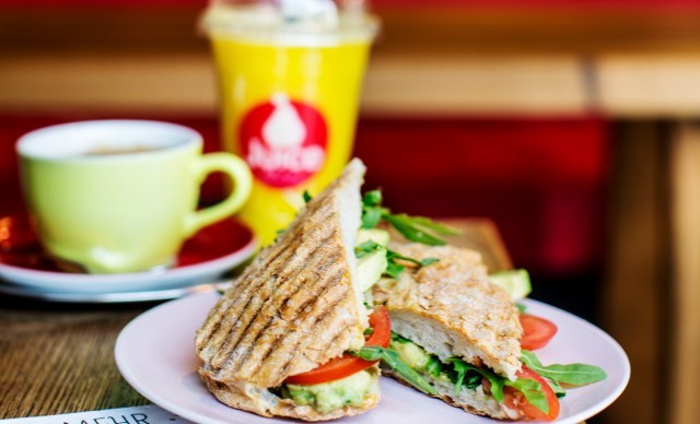 Ein Panini-Sandwich mit Tomaten, Avocado und Rucola auf einem Teller, daneben ein gelber Smoothie und eine Tasse Kaffee.