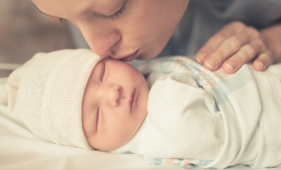 Mutter küsst ihr neugeborenes Baby auf die Stirn, das friedlich in eine Decke gewickelt schläft.