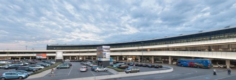 Panoramaaufnahme des Vorplatzes vom Flughafen Wien mit Parkplätzen, Autos und Bussen.