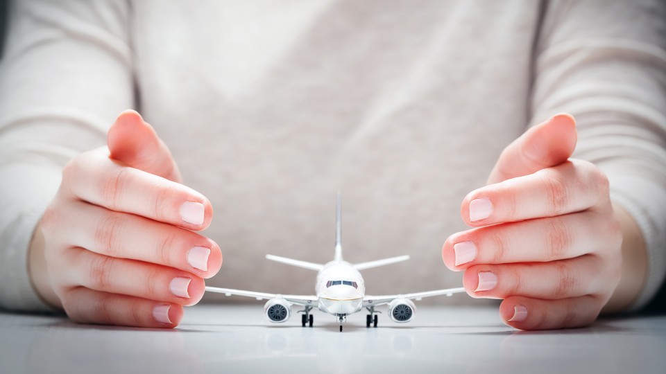 Zwei schützende Hände umschließen ein Modellflugzeug auf einem Tisch – Symbol für Sicherheit oder Schutz im Luftverkehr.