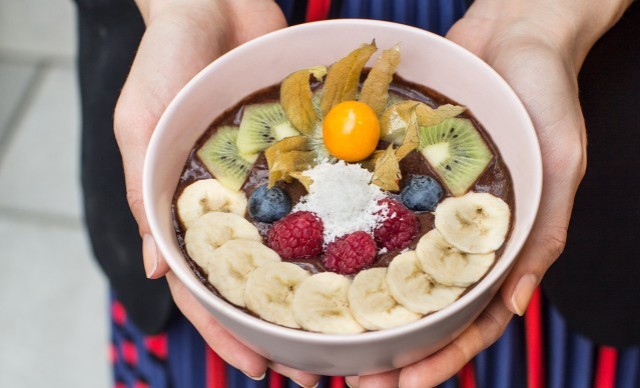 Hände halten eine Schüssel mit Smoothie-Bowl, dekoriert mit Bananenscheiben, Himbeeren, Blaubeeren, Kiwi und Physalis