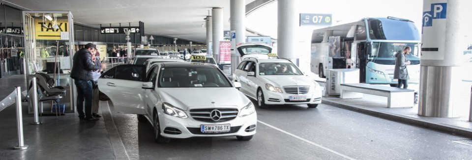 Taxistand am Flughafen mit wartenden Taxis und Fahrgästen, Reisebus im Hintergrund.