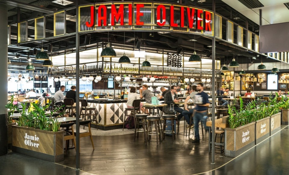 Innenansicht des Jamie Oliver Restaurants am Flughafen Wien mit modernem Design, offener Bar, grünen Hängelampen und Gästen an Tischen.