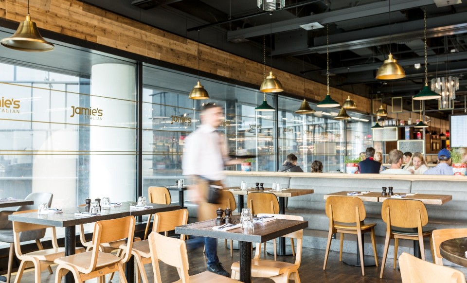 Helles Restaurant mit Holzmöbeln, leeren Tischen im Vordergrund und Gästen im Hintergrund. Ein Mann geht durch das Bild .
