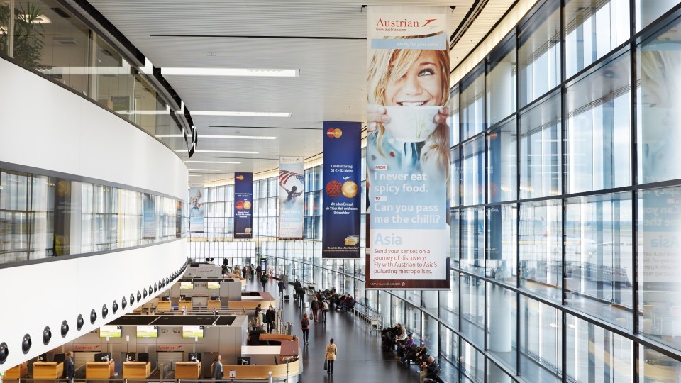 Helle Abflughalle am Flughafen Wien mit großen Panoramafenstern, Check-in-Schaltern von Austrian Airlines im Vordergrund und hängenden Werbebannern, die asiatische Reiseziele bewerben. Viele Reisende bewegen sich durch die Halle.