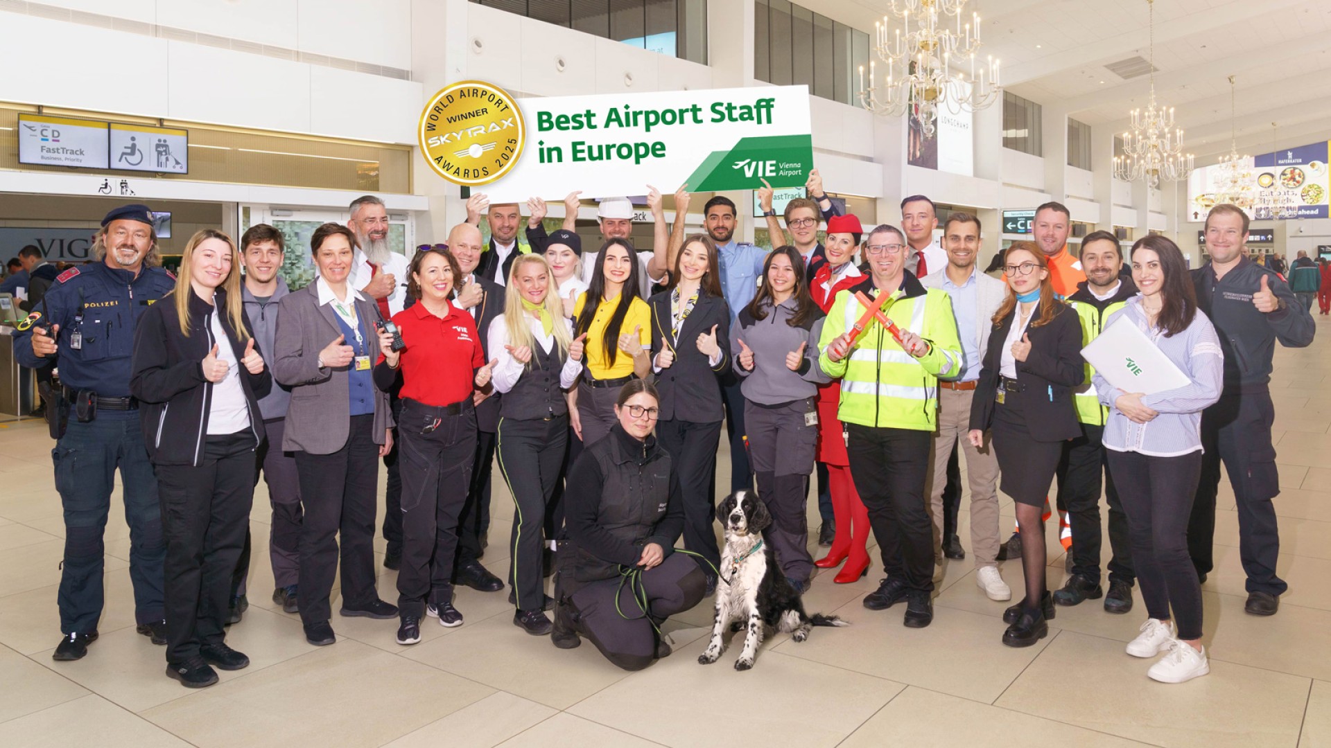 Group of airport staff in uniform posing in the departure hall of Vienna Airport with a sign reading: 'Best Airport Staff in Europe'.