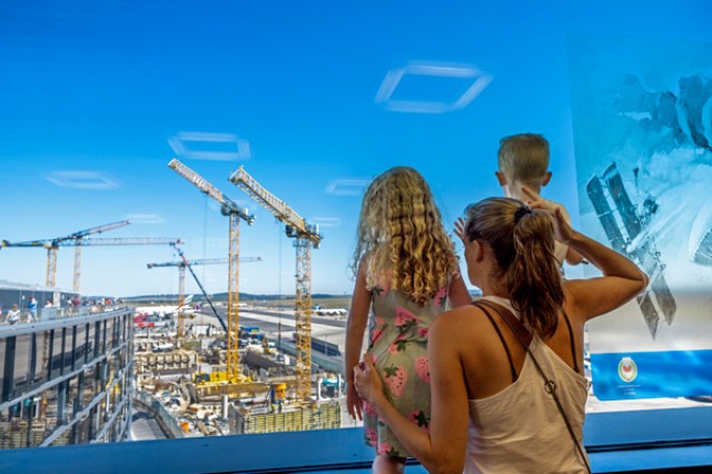 Mutter mit Kindern blickt auf Bauarbeiten am Flughafen durch Glaswand.