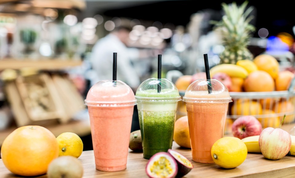 Drei Smoothies in Plastikbechern mit Strohhalmen, umgeben von verschiedenen Früchten wie Ananas, Äpfeln und Passionsfrüchten .