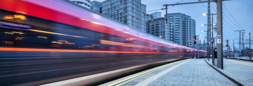Schnell fahrender Zug bei der Einfahrt in einen modernen Bahnhof, Hochhäuser im Hintergrund.