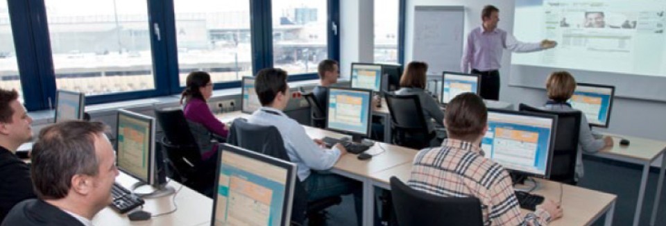 Ein Schulungsraum mit mehreren Personen, die an Computern arbeiten. Ein Trainer steht vorne und erklärt Inhalte, die auf eine Leinwand projiziert werden. Große Fenster bieten Ausblick auf den Flughafen.