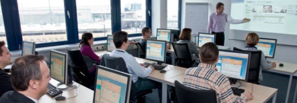 A training room with several people working on computers. A trainer stands at the front explaining content projected onto a screen. Large windows offer a view of the airport.