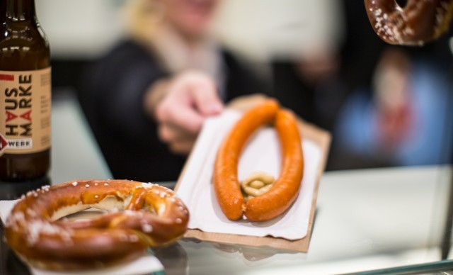 Zwei Brezeln und eine Flasche Bier auf einem Tisch, im Hintergrund reicht eine Person eine Wurst.