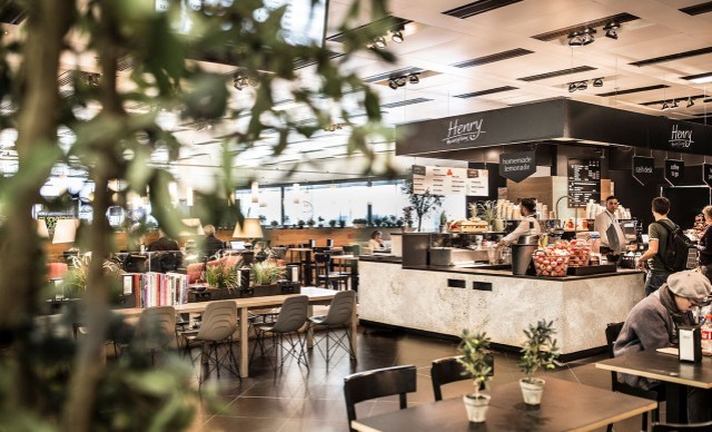Modernes Café mit Theke, Sitzbereich und Pflanzen im Vordergrund. Menschen bestellen und genießen Getränke.