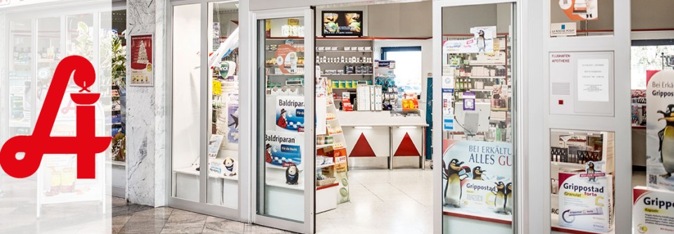 Eingang einer Apotheke mit Regalen voller Medikamente und Gesundheitsprodukte, Werbeplakate für Grippostad und andere Produkte.