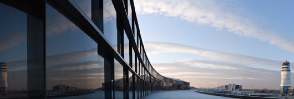 Außenansicht des Terminal 3 am Flughafen Wien mit reflektierenden Glasfassaden, die die Struktur eines langgezogenen Gebäudes zeigen. Im Vordergrund spiegeln sich Wolken und Himmel in den Fenstern, während im Hintergrund der charakteristische Kontrollturm des Flughafens zu sehen ist.