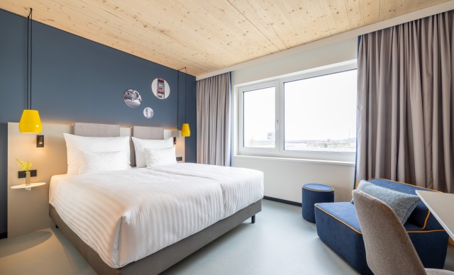 Modernes Hotelzimmer mit einem Doppelbett, großem Fenster, blauen Sesseln und einem Schreibtisch. Über dem Bett hängen drei runde Wandbilder, und gelbe Pendelleuchten beleuchten den Schlafbereich.