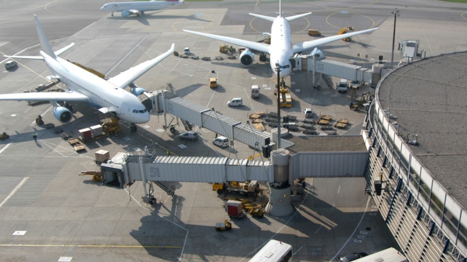 Zwei große Passagierflugzeuge sind an Fluggastbrücken eines Terminals angedockt, rundherum befinden sich verschiedene Bodenfahrzeuge und Gepäckwagen.