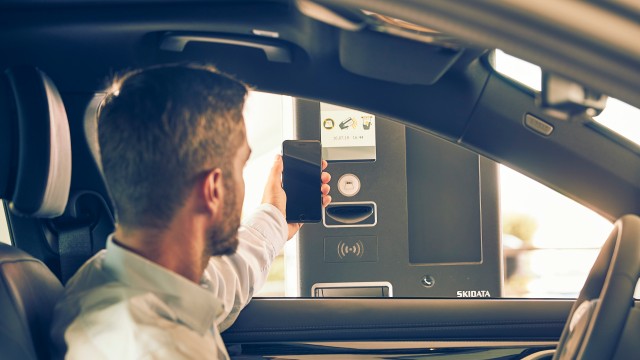 Ein Mann sitzt in einem Auto und hält sein Smartphone an eine Schrankenanlage, um kontaktlos zu bezahlen oder ein Parkticket zu scannen. Die Szene zeigt ein System von SKIDATA.