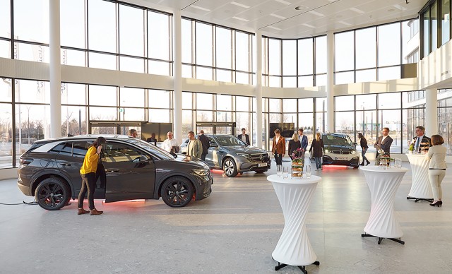 Automesse, mit drei schwarzen Autos in einer Halle und verschiedensten Personen, welche diese begutachten