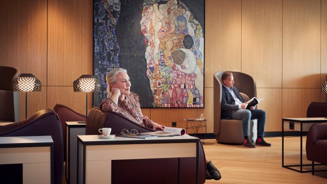 Zwei Personen entspannen sich in einer stilvollen Flughafenlounge mit warmem Holzinterieur. Im Vordergrund sitzt eine Frau mit Zeitung und Kaffeetasse, im Hintergrund liest ein Mann ein Buch. An der Wand hängt ein großes Gemälde im Stil von Gustav Klimt. Die Atmosphäre wirkt ruhig und komfortabel.