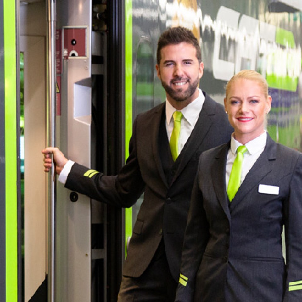 Zugbegleiter und Zugbegleiterin in Uniform mit grüner Krawatte stehen lächelnd am Einstieg eines modernen Zugs.