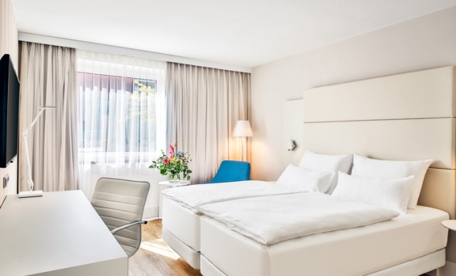 Hotelzimmer, ausgestattet mit einem großem, beigen Bett und einem weißen, großen Schreibtisch