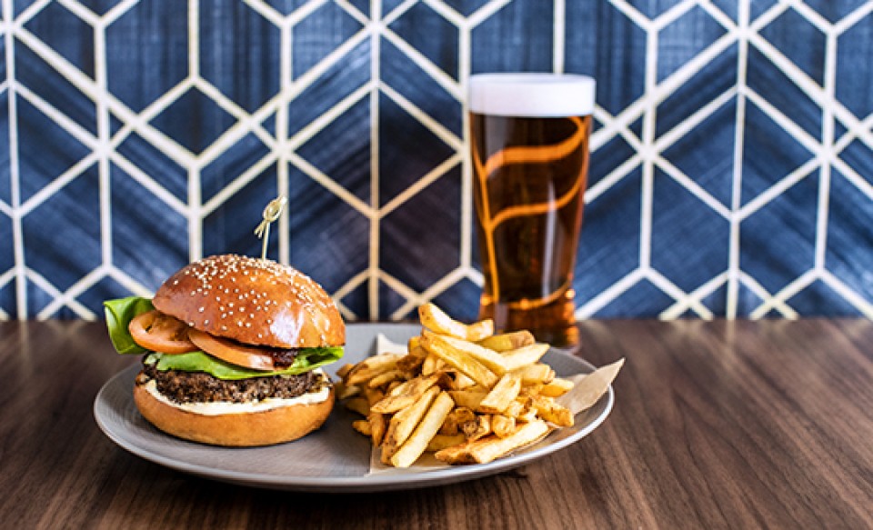 Saftiger Burger mit Pommes auf einem Teller, dazu ein Glas Bier – serviert vor geometrisch gemusterter Wand.