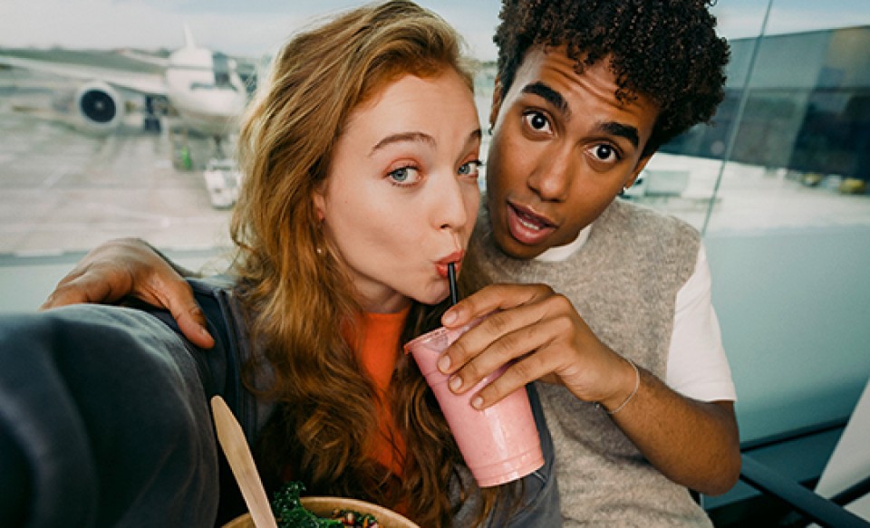 A young couple at an airport sharing a drink while taking a selfie, with an aircraft visible in the background.